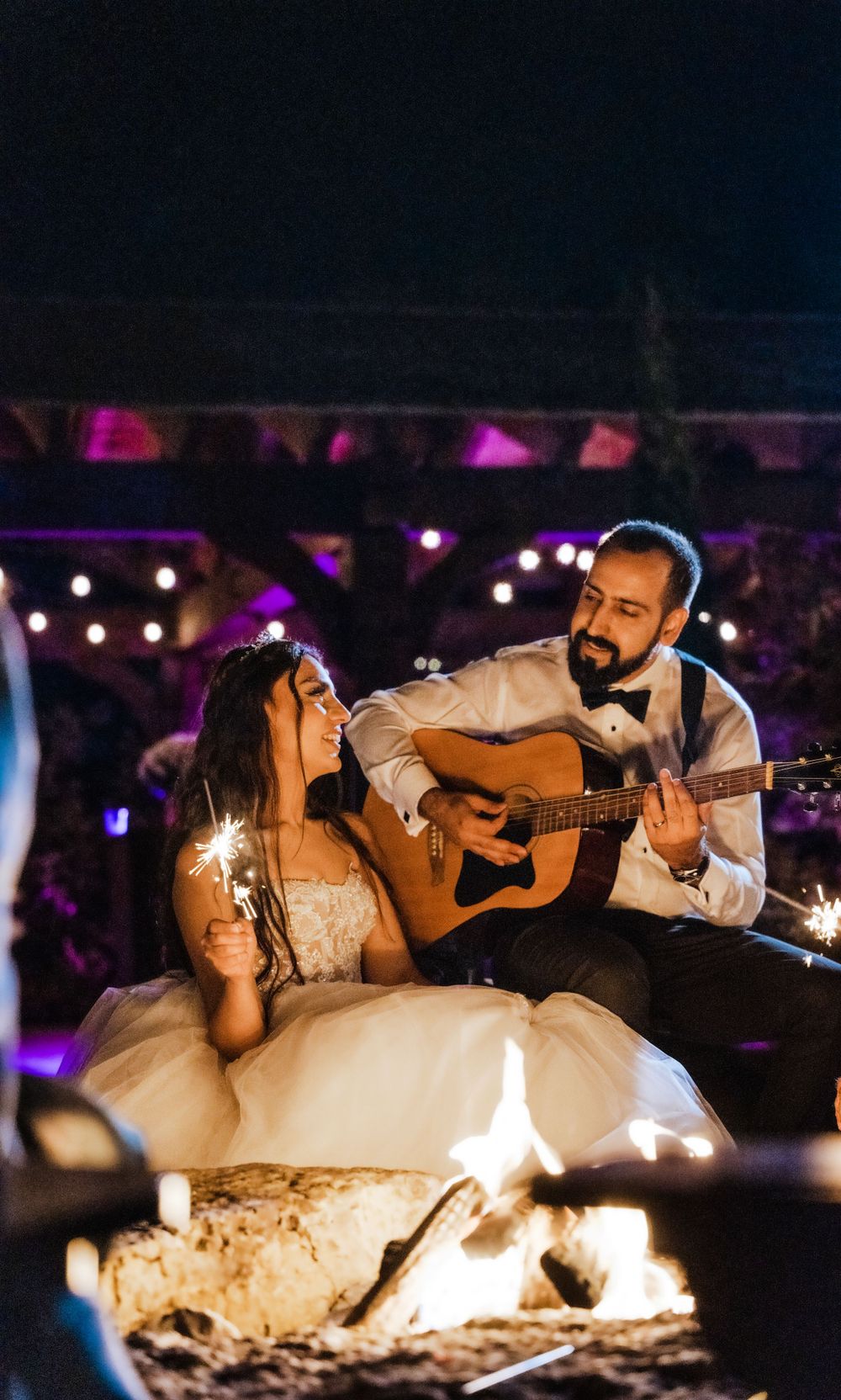 A couple shares an intimate moment by a campfire while the groom plays acoustic guitar.