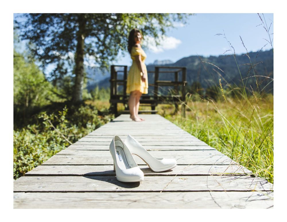 White high heels placed on a wooden boardwalk with a figure standing in a yellow dress in the background.