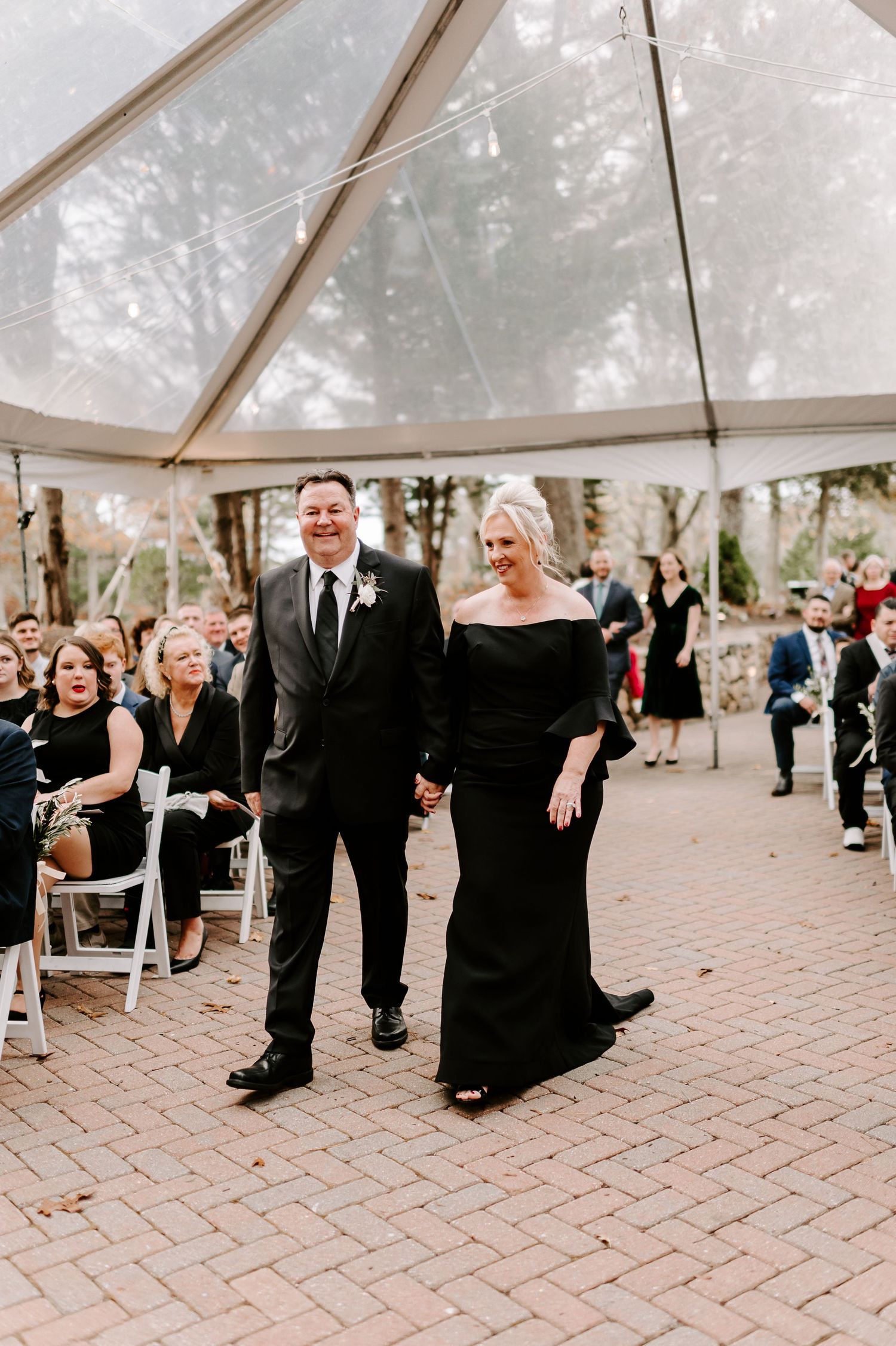 Wedding guests dancing under a clear tent at an outdoor reception on brick pavers.