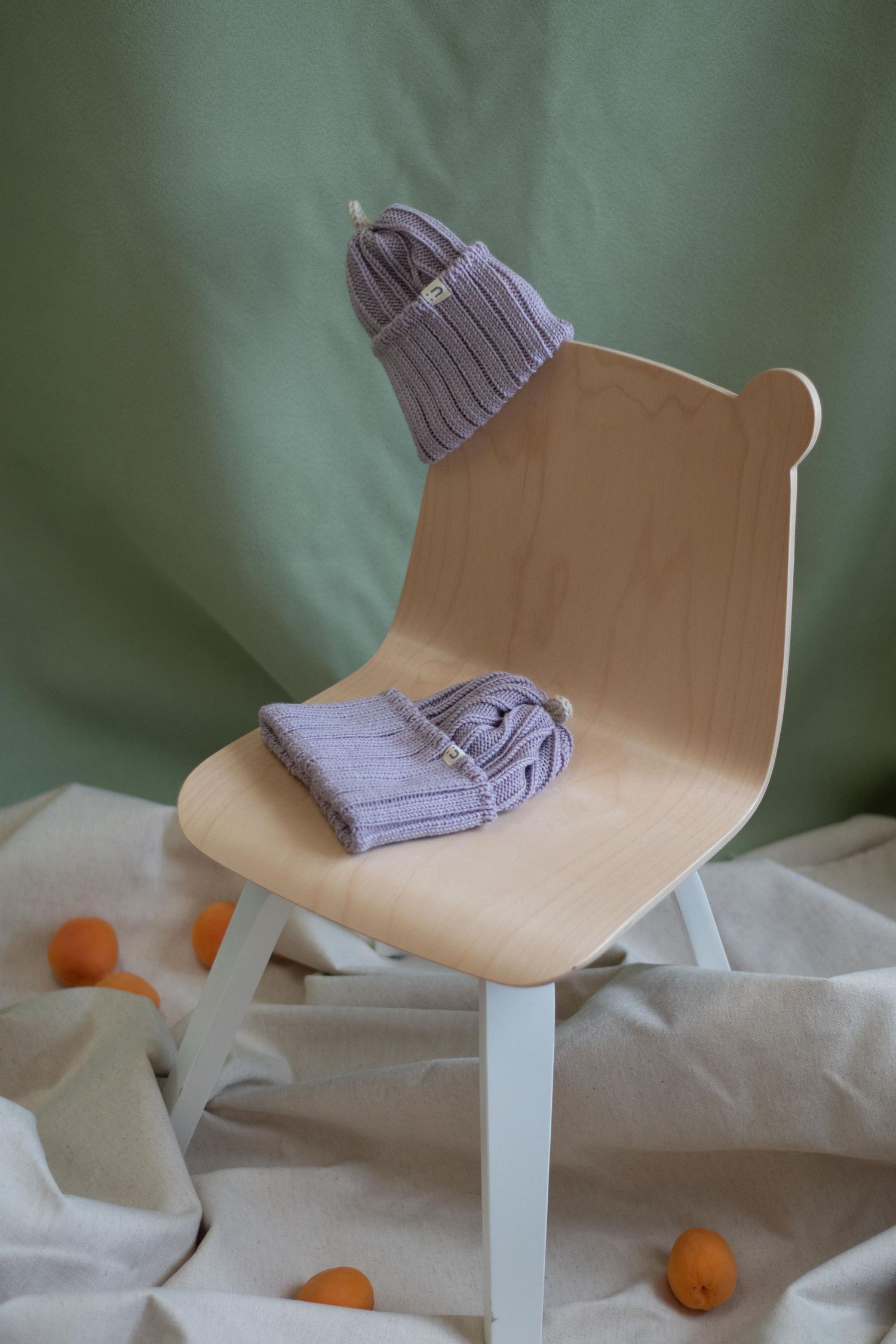 Lilac knit beanie displayed on minimalist wooden chair against sage green background.