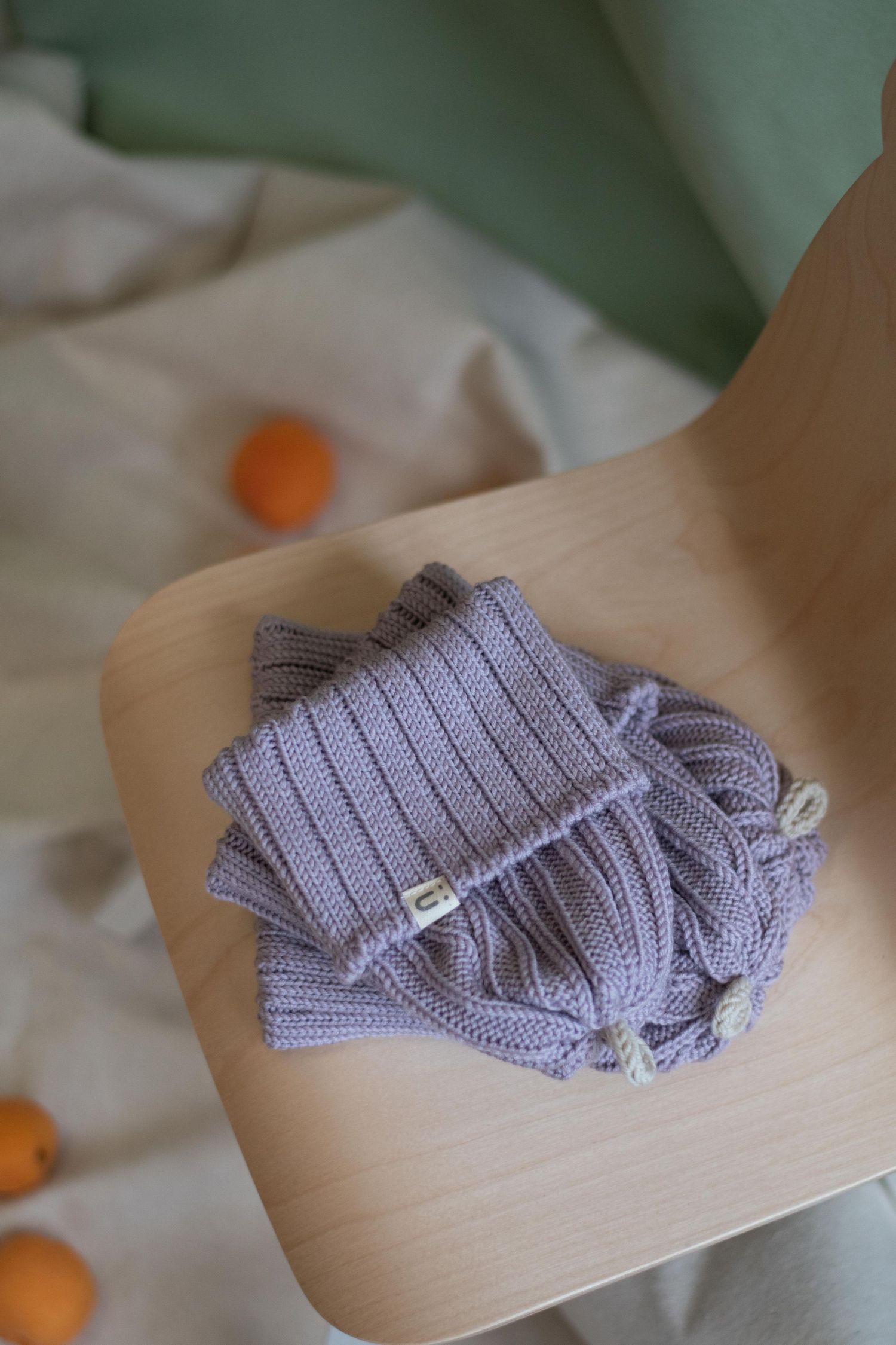 Folded lilac knit beanie on wooden chair with scattered oranges.