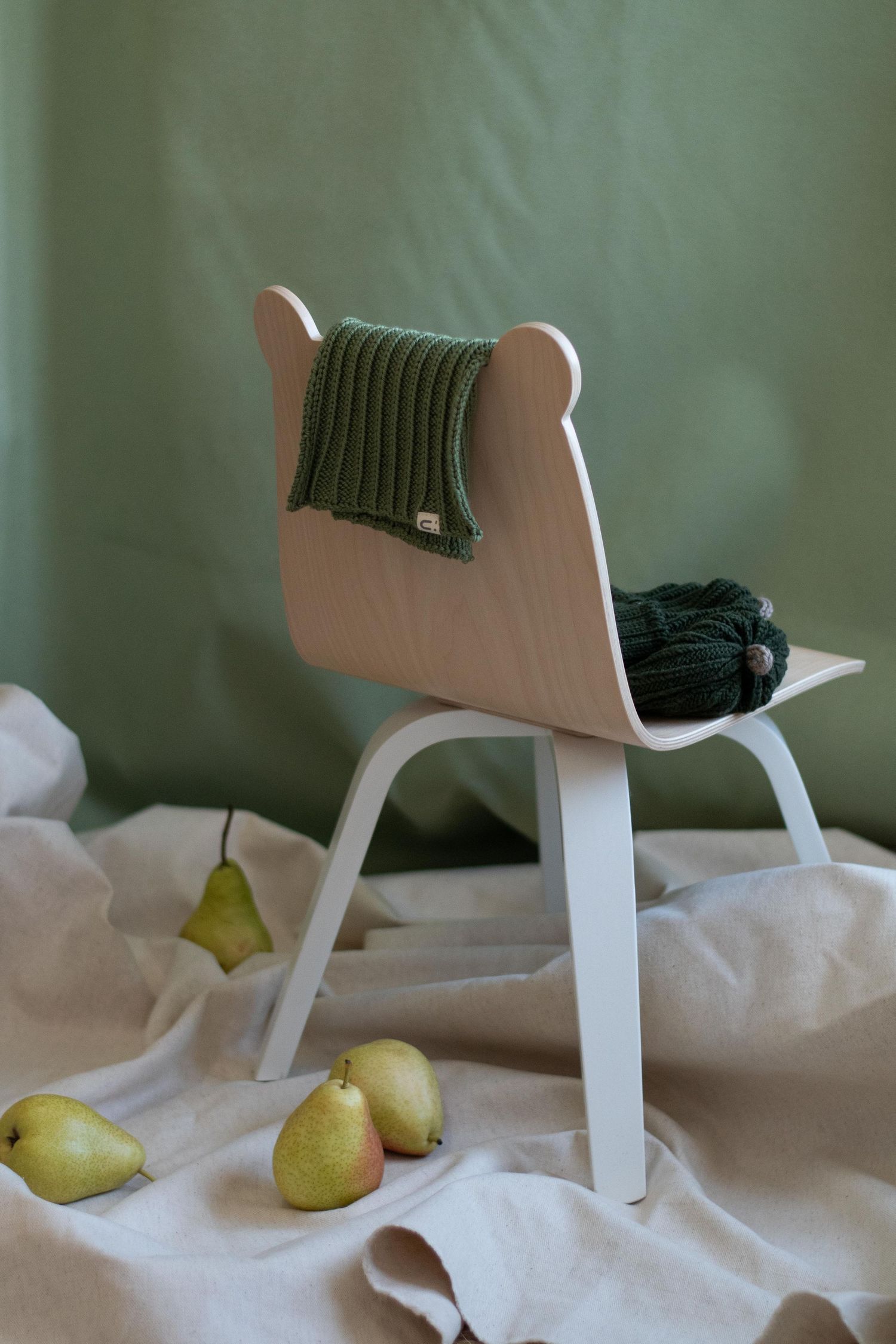 Modern wooden chair with dark green knit beanie and pears on white fabric.