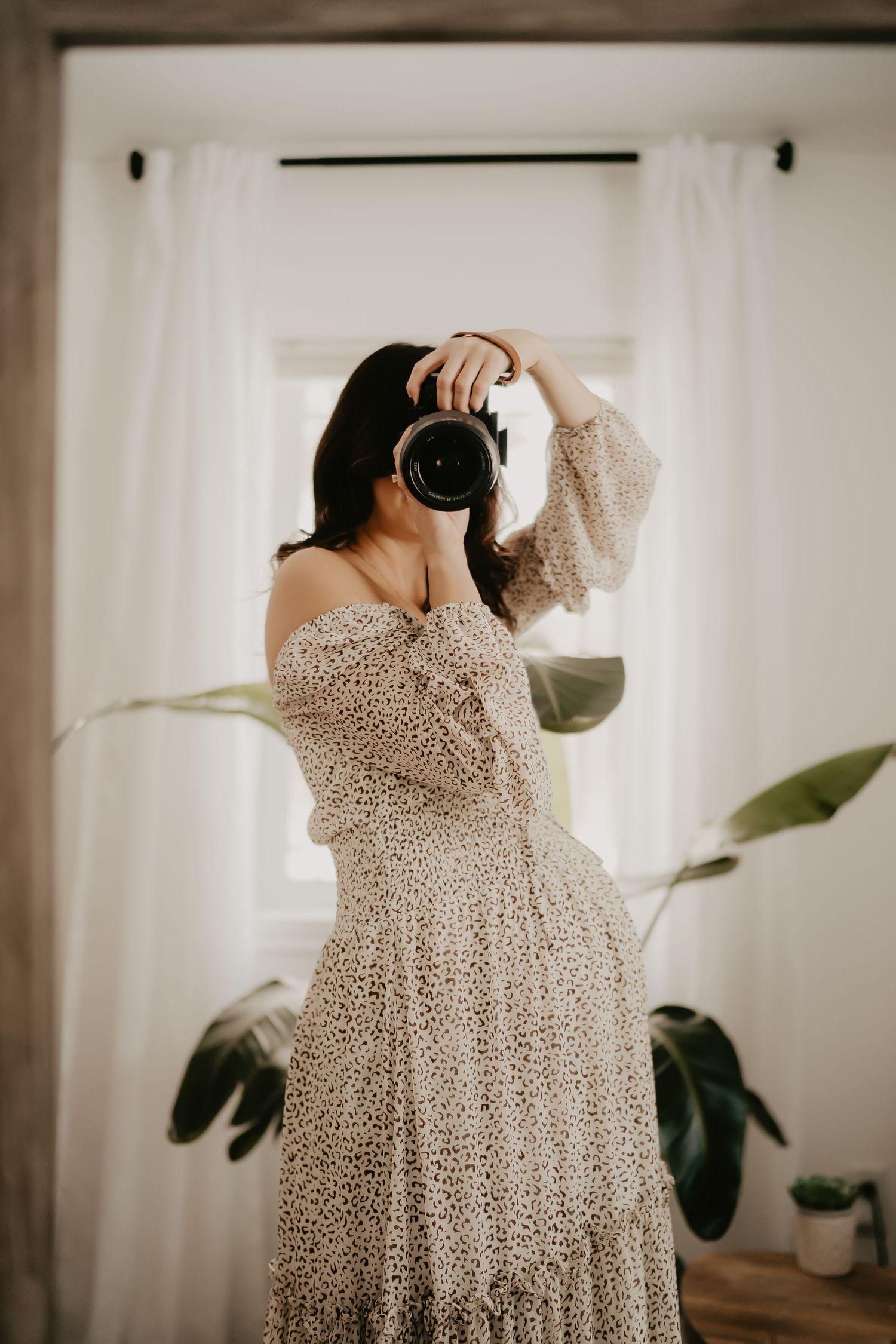 Photographer in a floral dress holds up camera against white curtains and indoor plants.