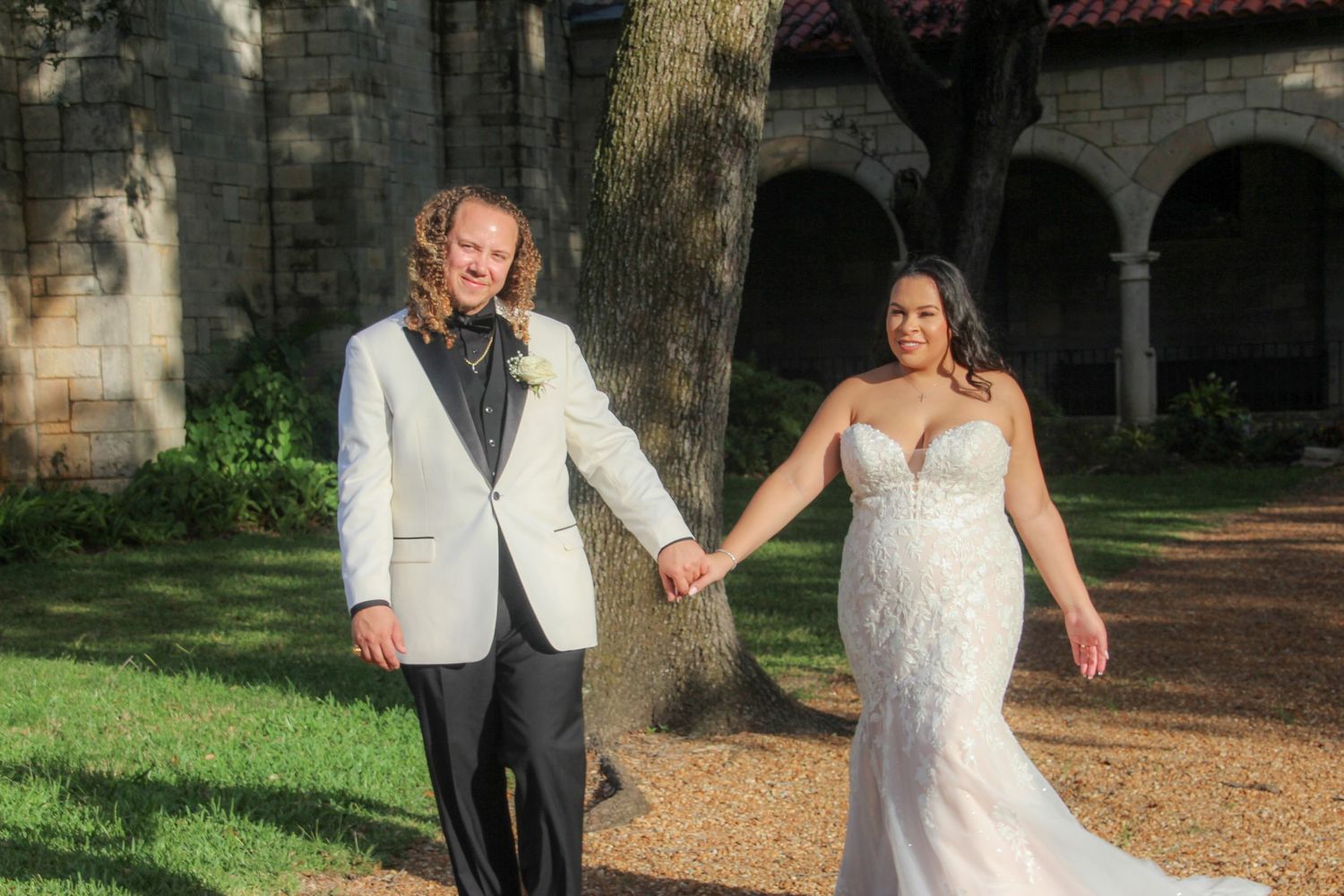 Bride in lace gown and groom in white jacket holding hands walking near stone building.