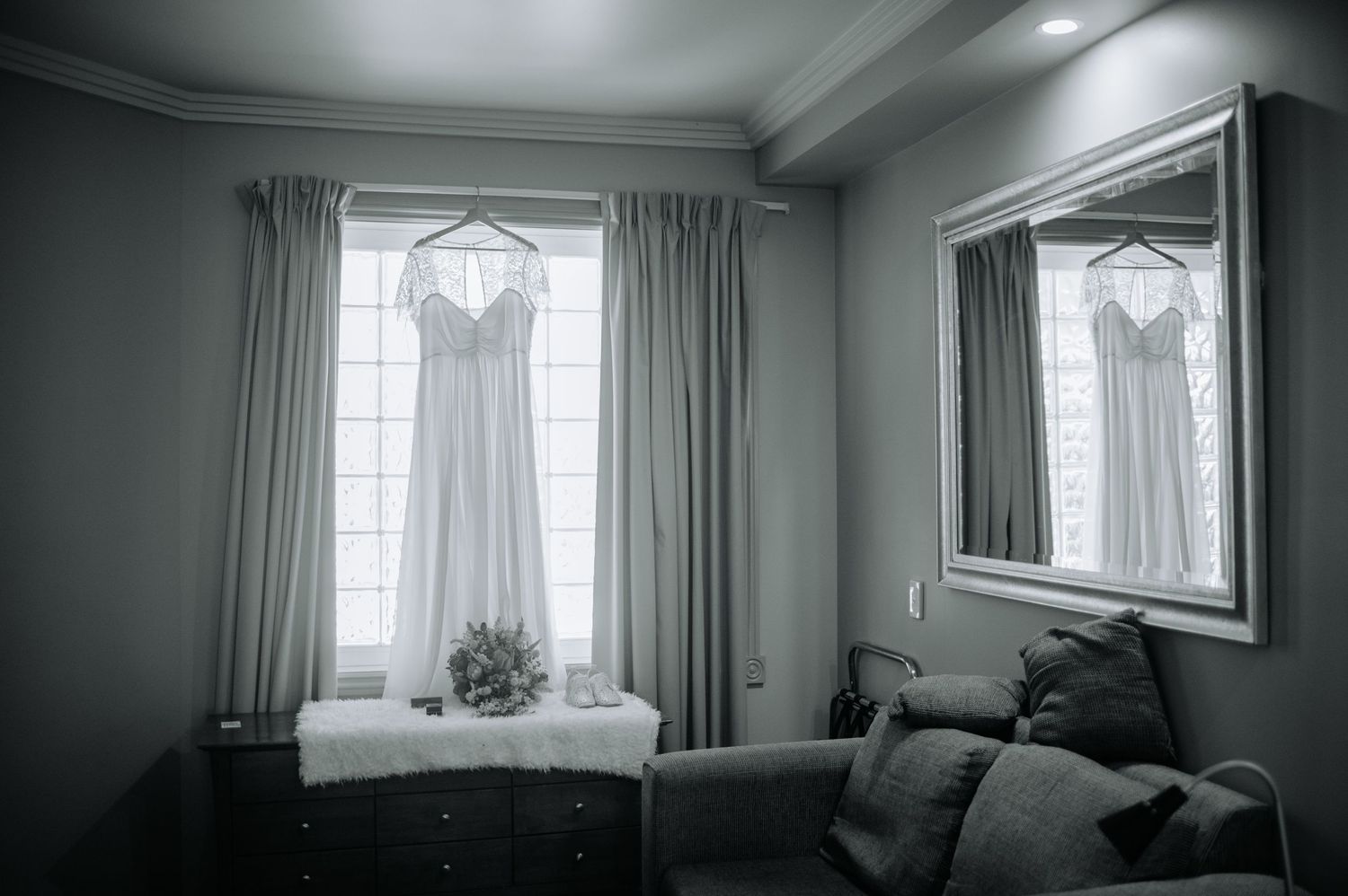 Wedding dress hangs in window light creating dramatic silhouette against grey walls.