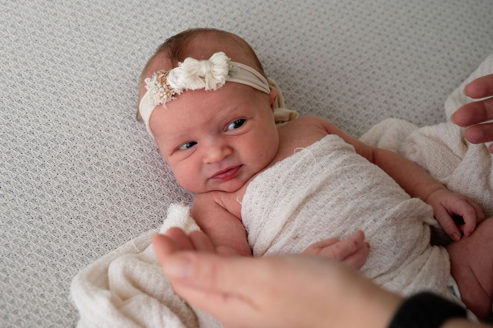 A swaddled newborn wearing a lace headband lies on soft grey fabric during a photo session.