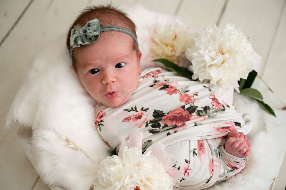 A small baby wearing floral print and headband poses with white peonies on plush fabric.