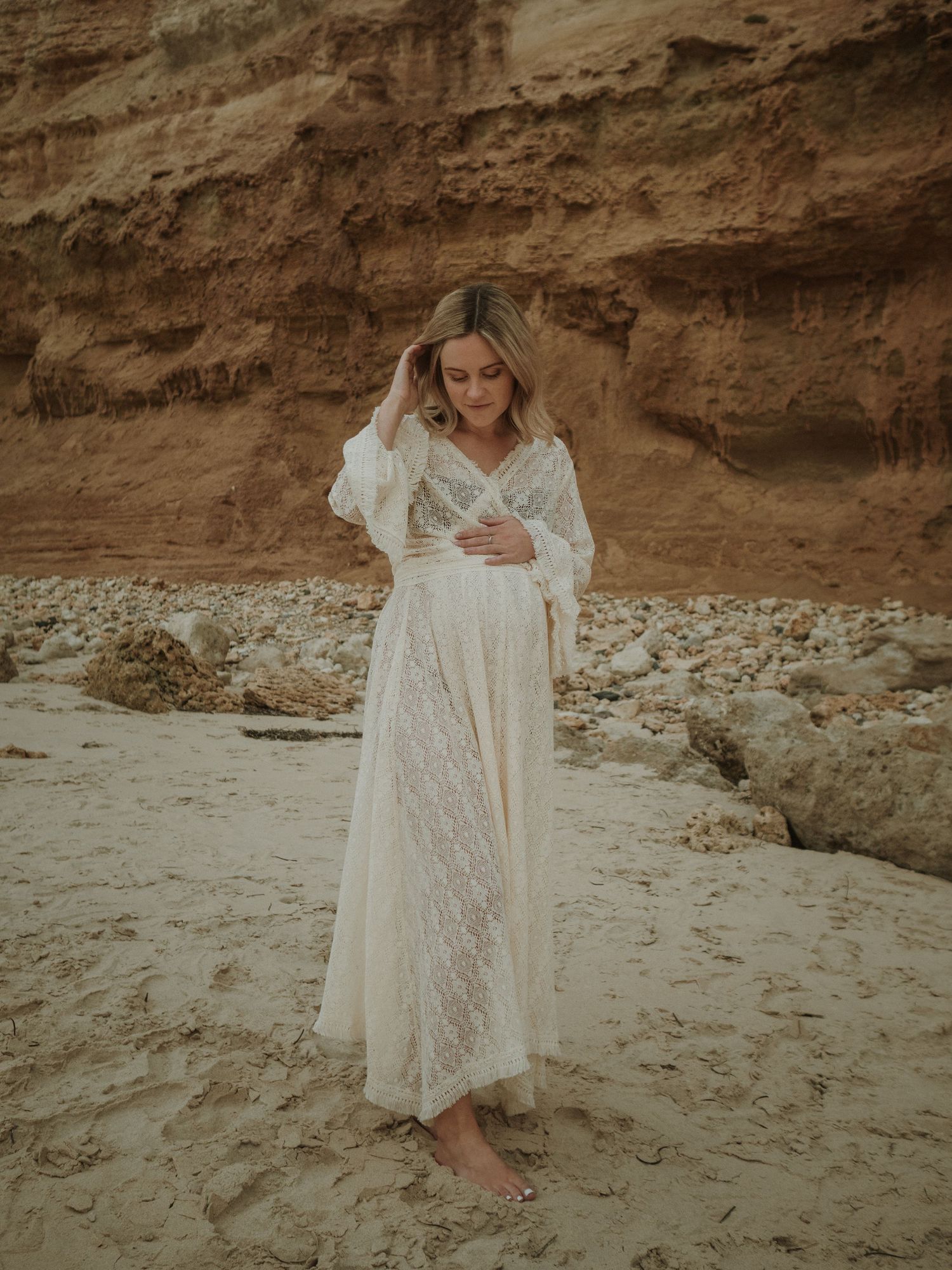 A white lace maternity dress stands out against dramatic coastal cliffs and sandy beach at sunset.