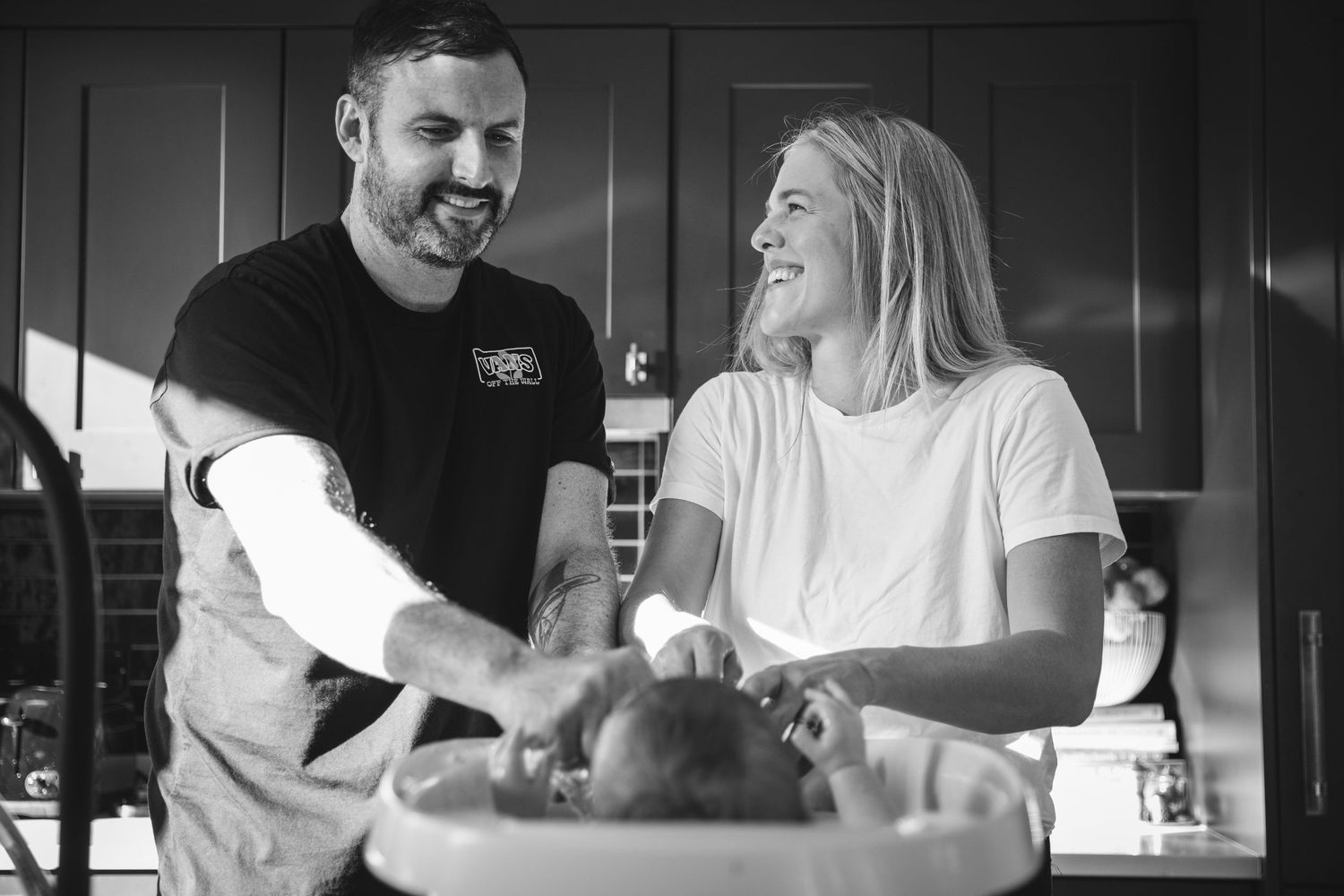 Candid black and white photo of couple laughing while cooking together in kitchen.