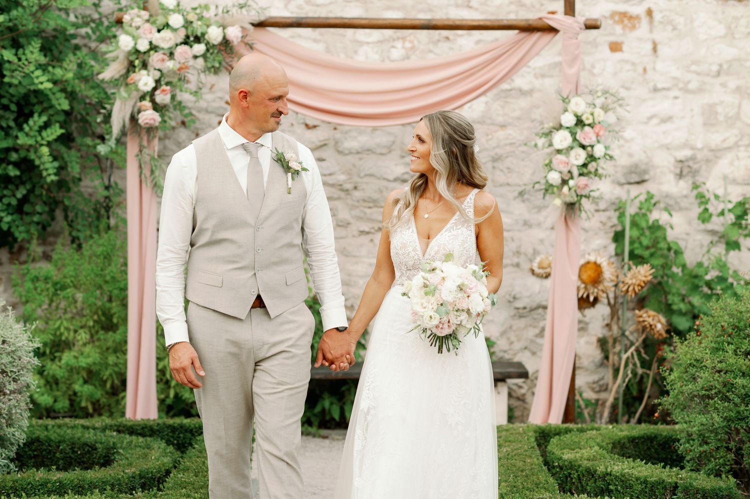Newlyweds hold hands and smile under a blush pink wedding arch with floral decorations.