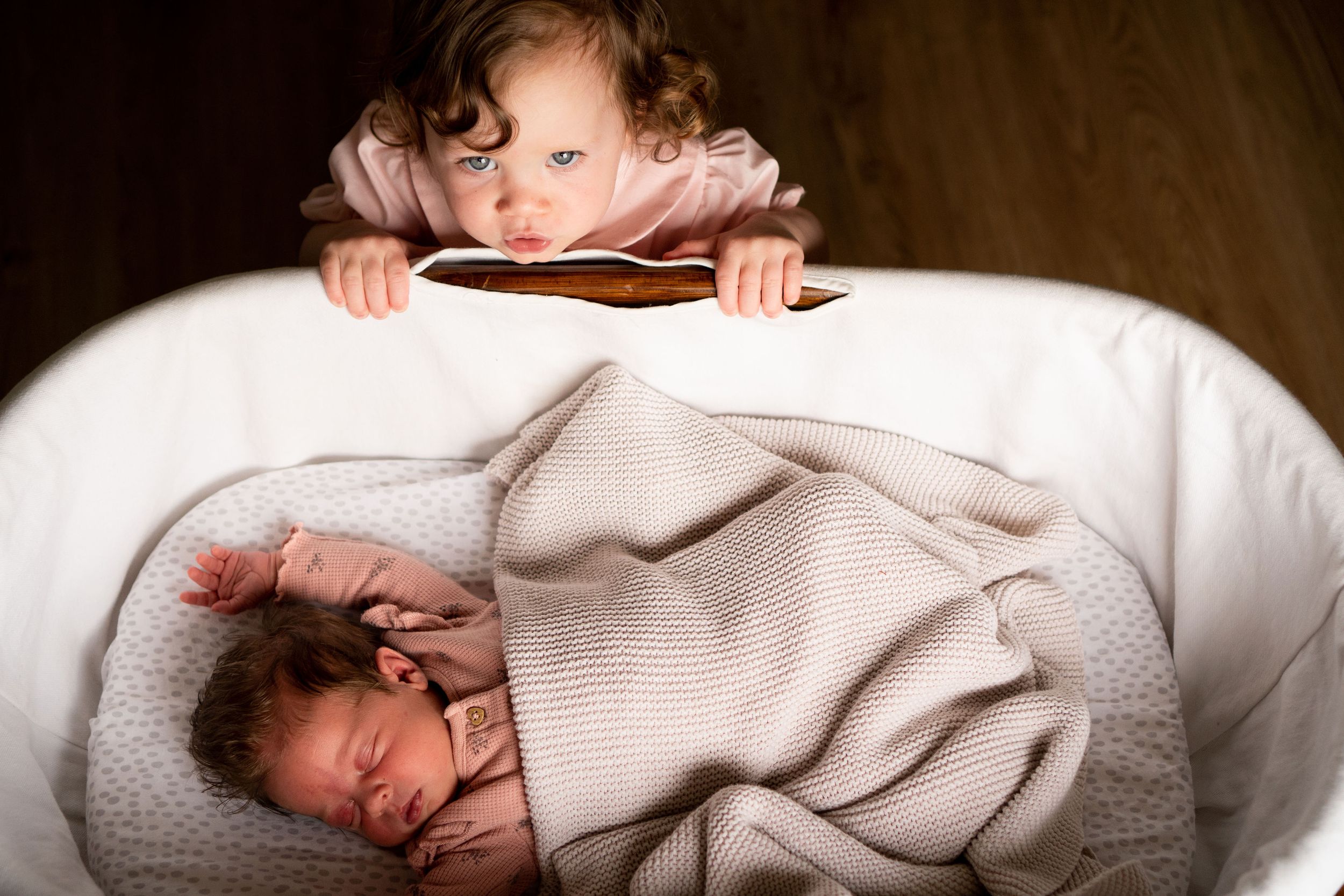 Newborn baby lying asleep in a bassinet, while older sister peaks over the side
