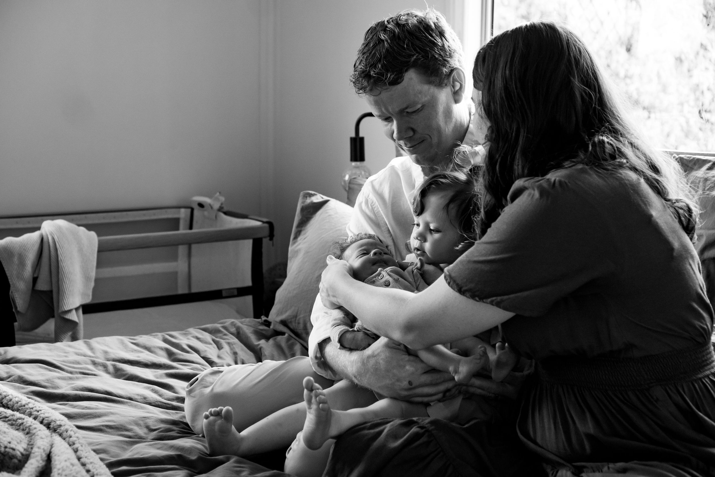 Mother and father seated on a bed, helping toddler to hold her newborn baby sister
