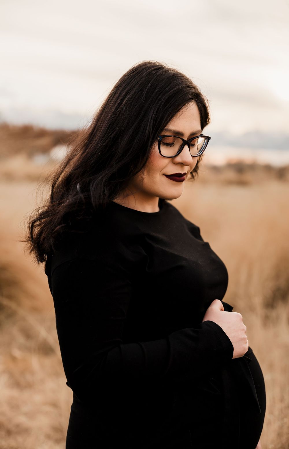 Maternity portrait in black dress and glasses against autumn landscape.