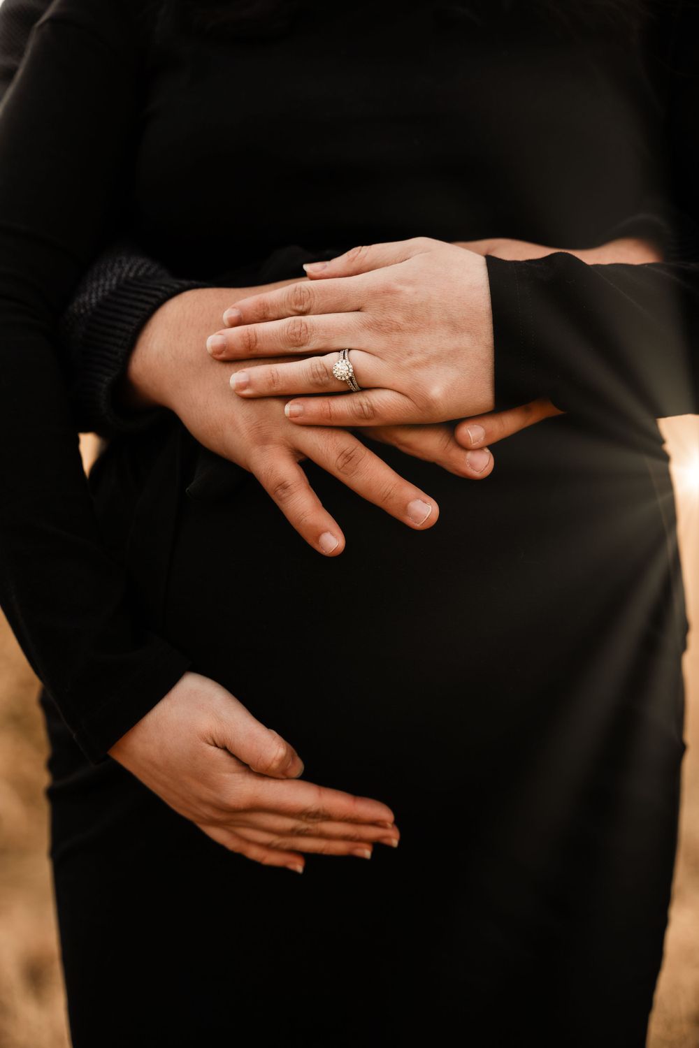 Close-up of hands with engagement ring against black clothing in warm sunlight.