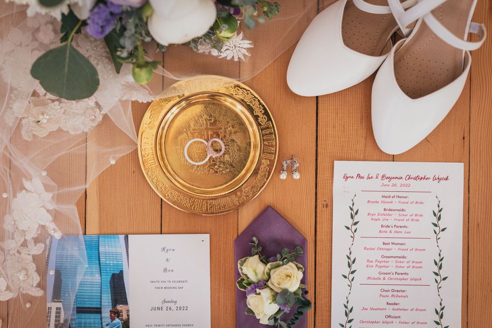 Wedding details flatlay featuring gold plate, white shoes, invitation and purple floral accents.
