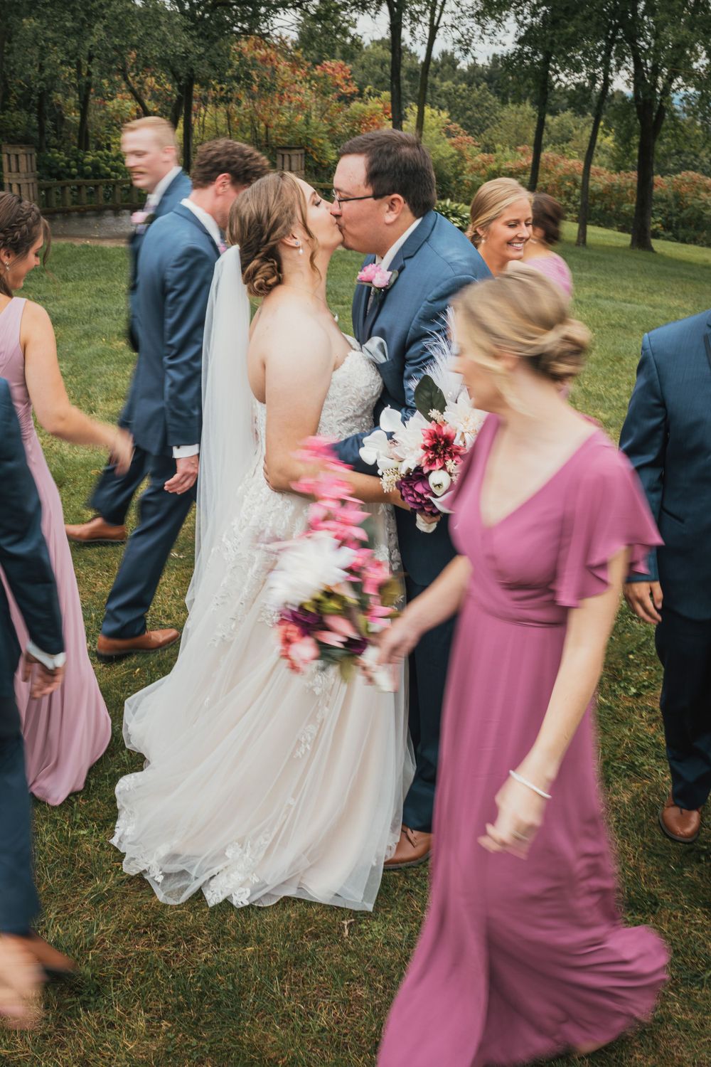 Wedding party walks outdoors with bridesmaids wearing mauve dresses holding colorful bouquets.