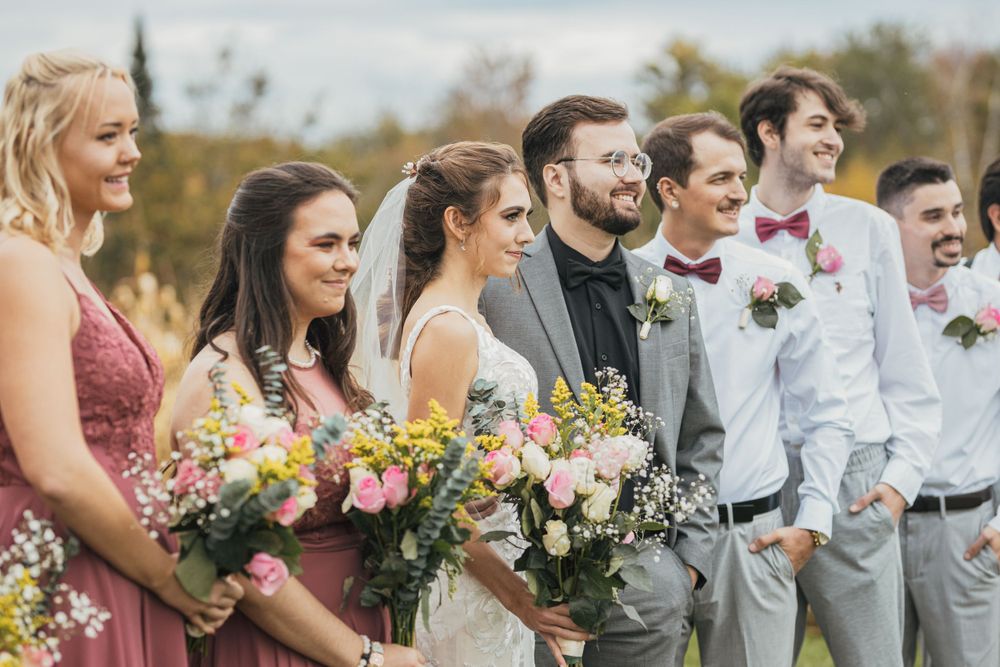 Wedding party standing together outdoors with bridesmaids in pink dresses and groomsmen in gray suits.