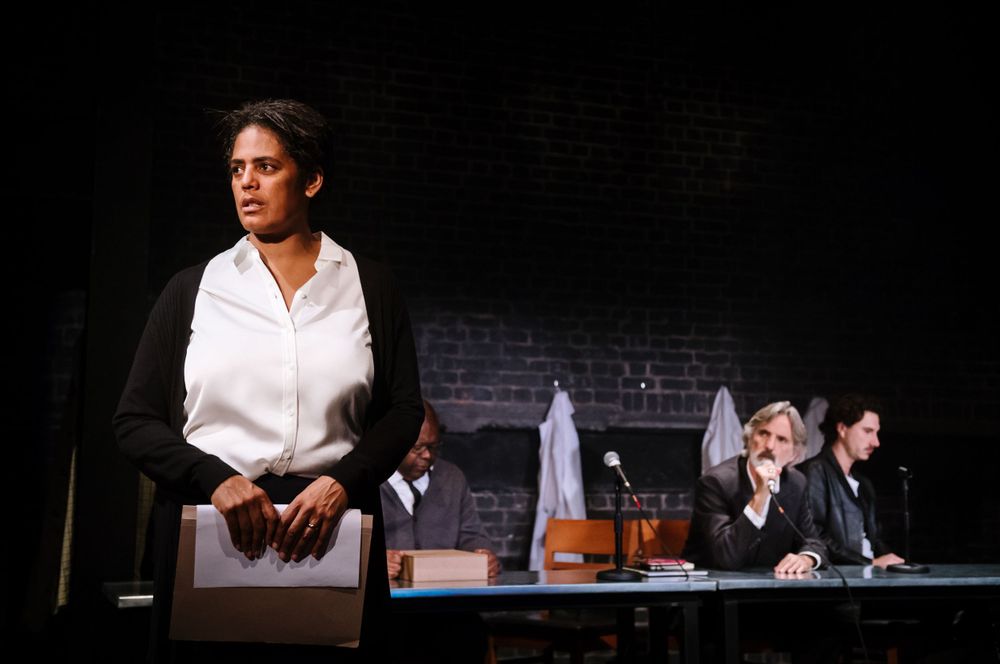 Theater scene with performers at office desk against dark brick wall backdrop.