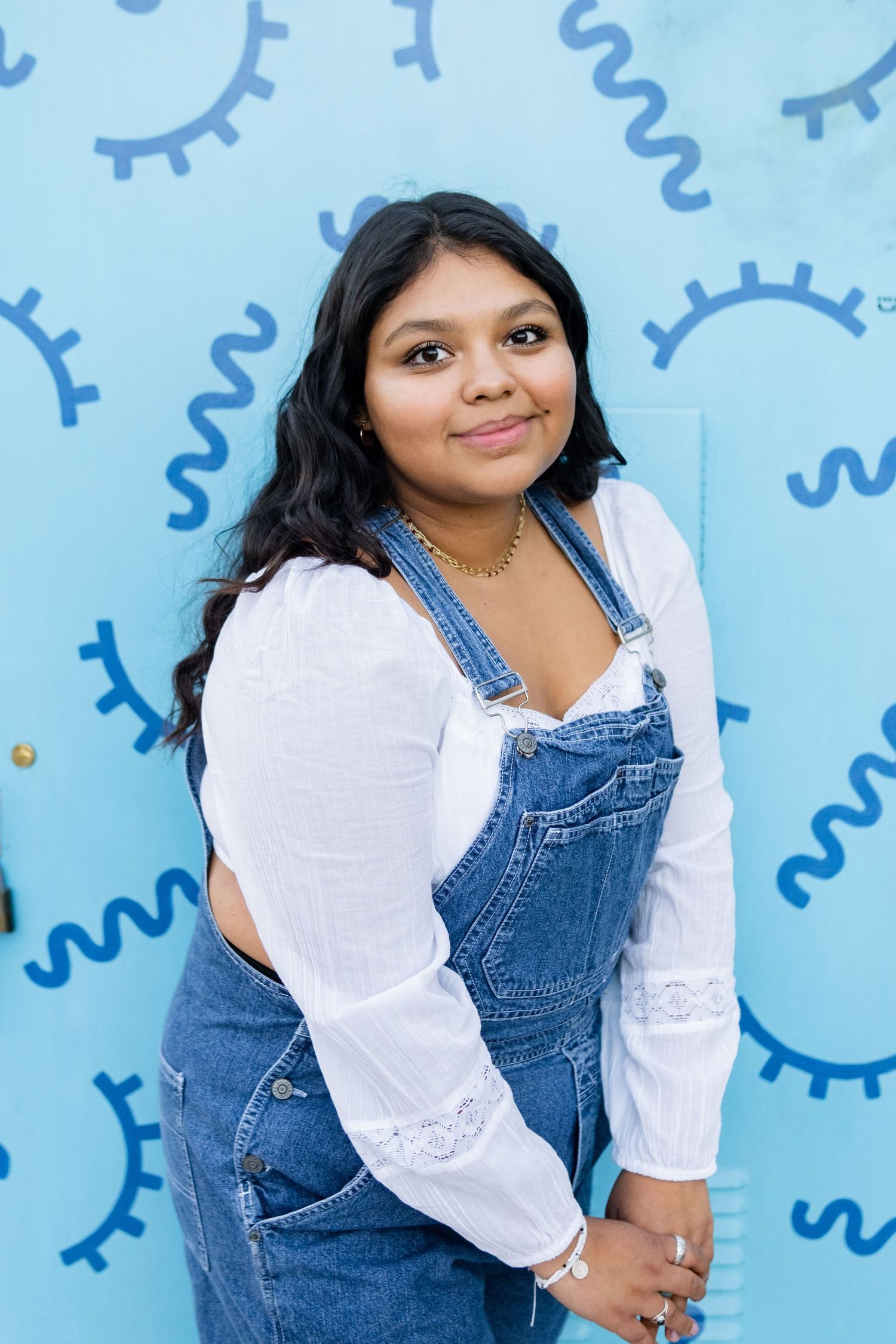 A high school senior in denim overalls and white blouse stands smiling against blue wall with whimsical gear pattern background.