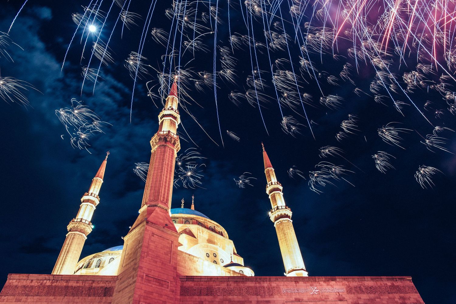 Beautiful mosque with three minarets illuminated at night with fireworks bursting in the dark blue sky.