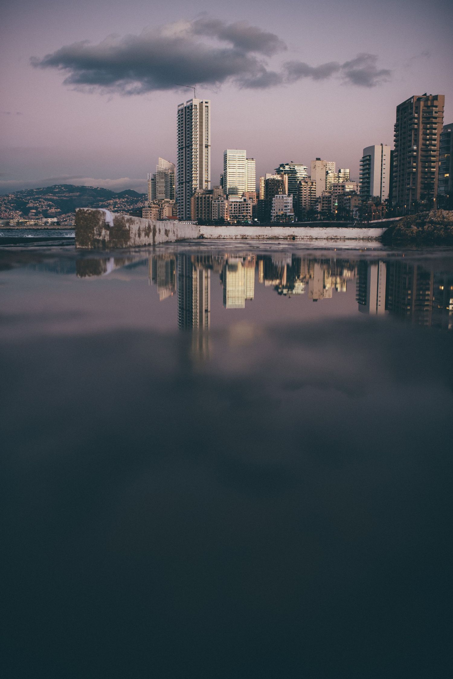 City skyline reflects in calm water during twilight with moody clouds.