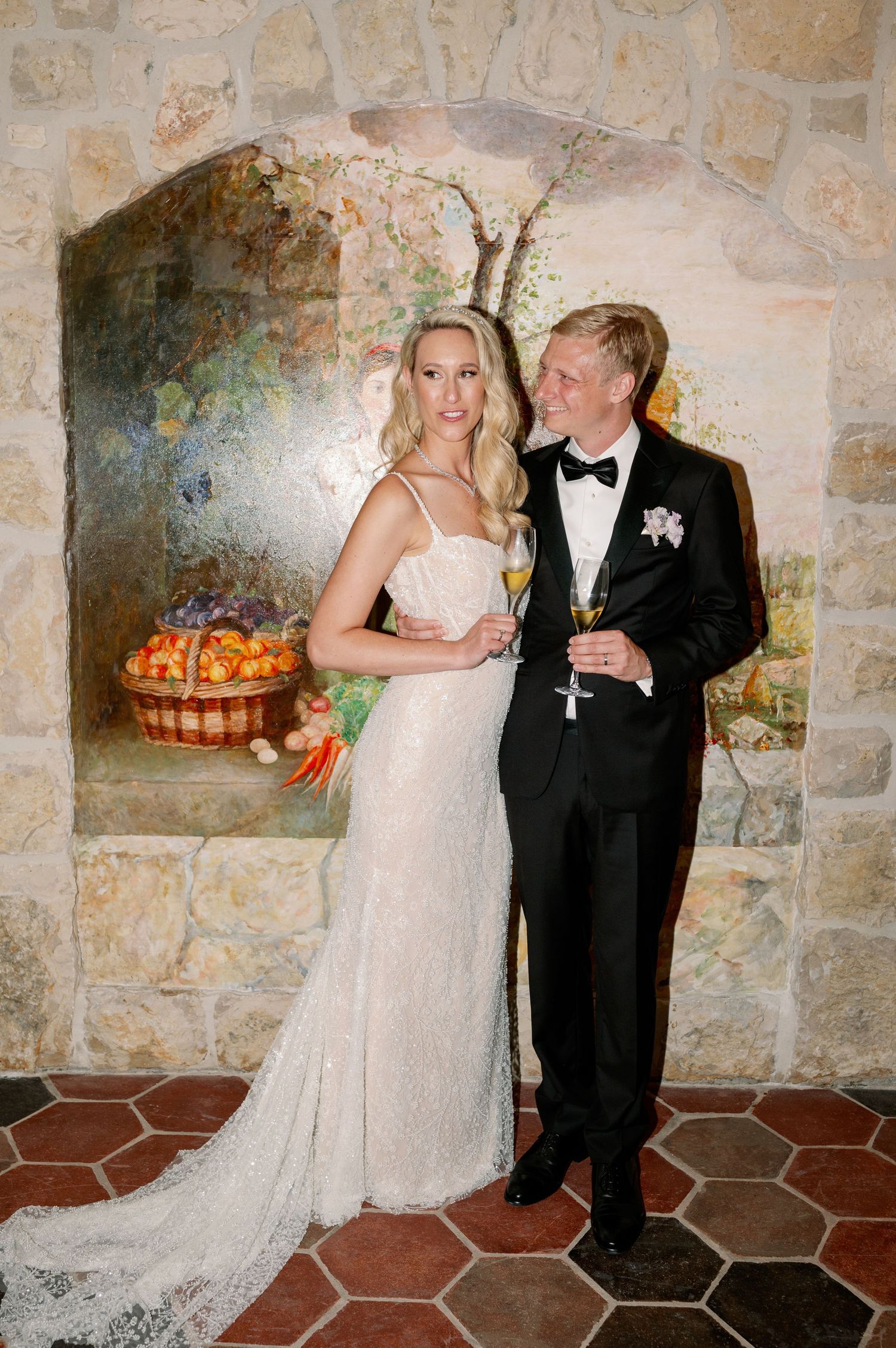 Wedding couple poses with champagne against rustic stone wall backdrop.