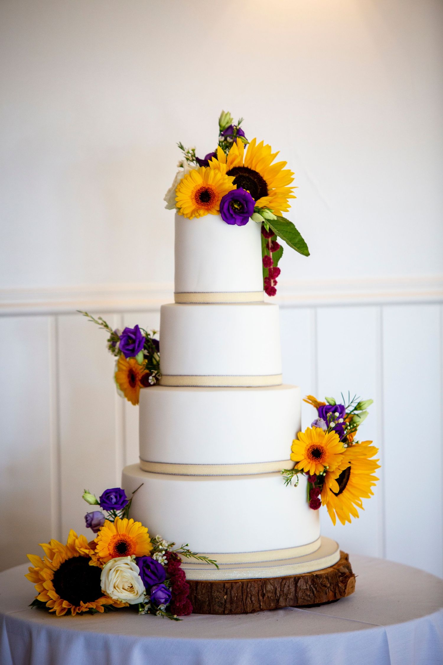 White tiered wedding cake decorated with sunflowers and purple flowers on a rustic wood base.
