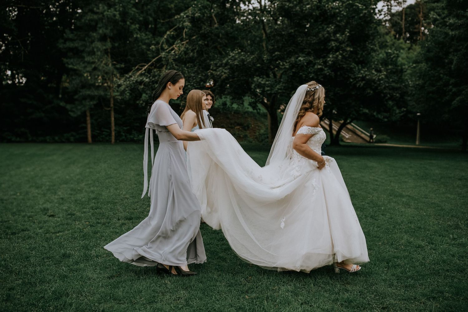 Three figures in flowing white dresses walk across a lush green lawn during a wedding celebration.