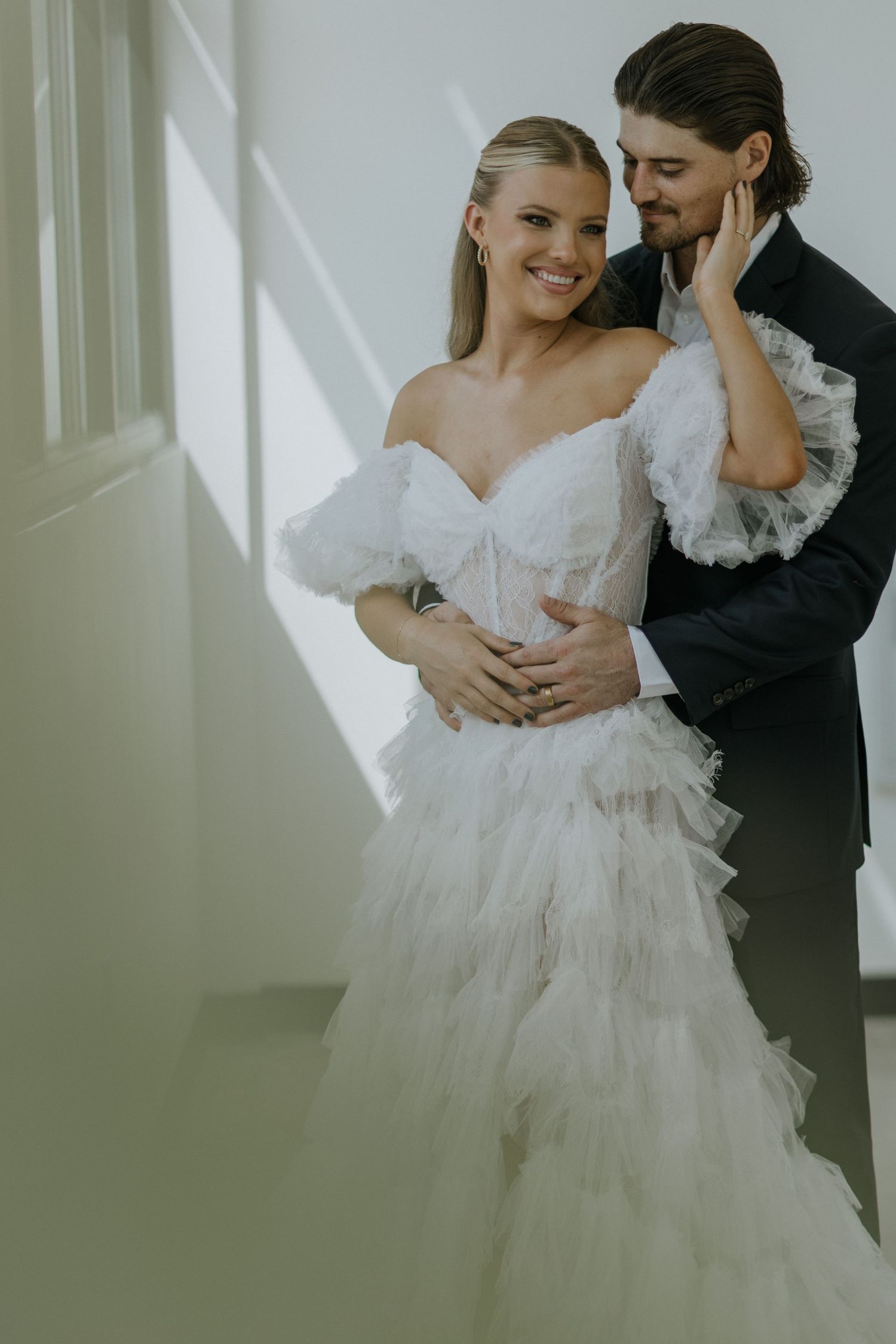 Elegant couple embracing in sunlit interior with flowing white wedding dress.