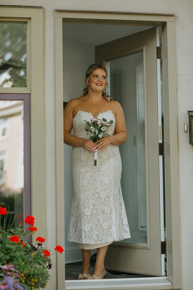 A joyful person in a white lace wedding dress stands in a doorway holding a small bouquet with red flowers in the garden.