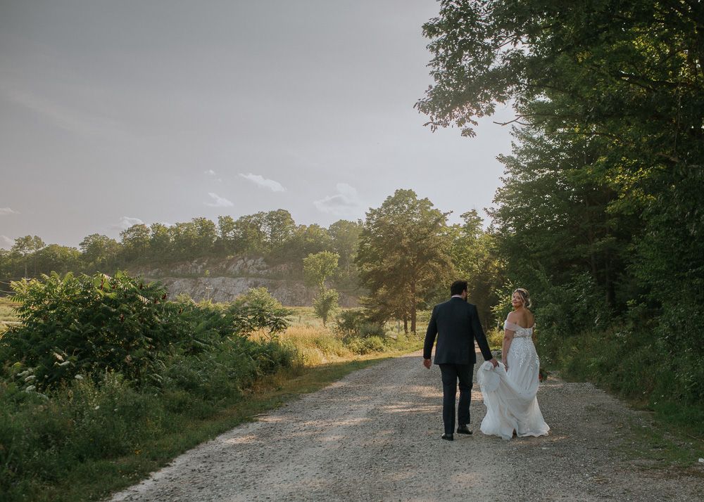Newlyweds walk hand in hand down a scenic country path at sunset.
