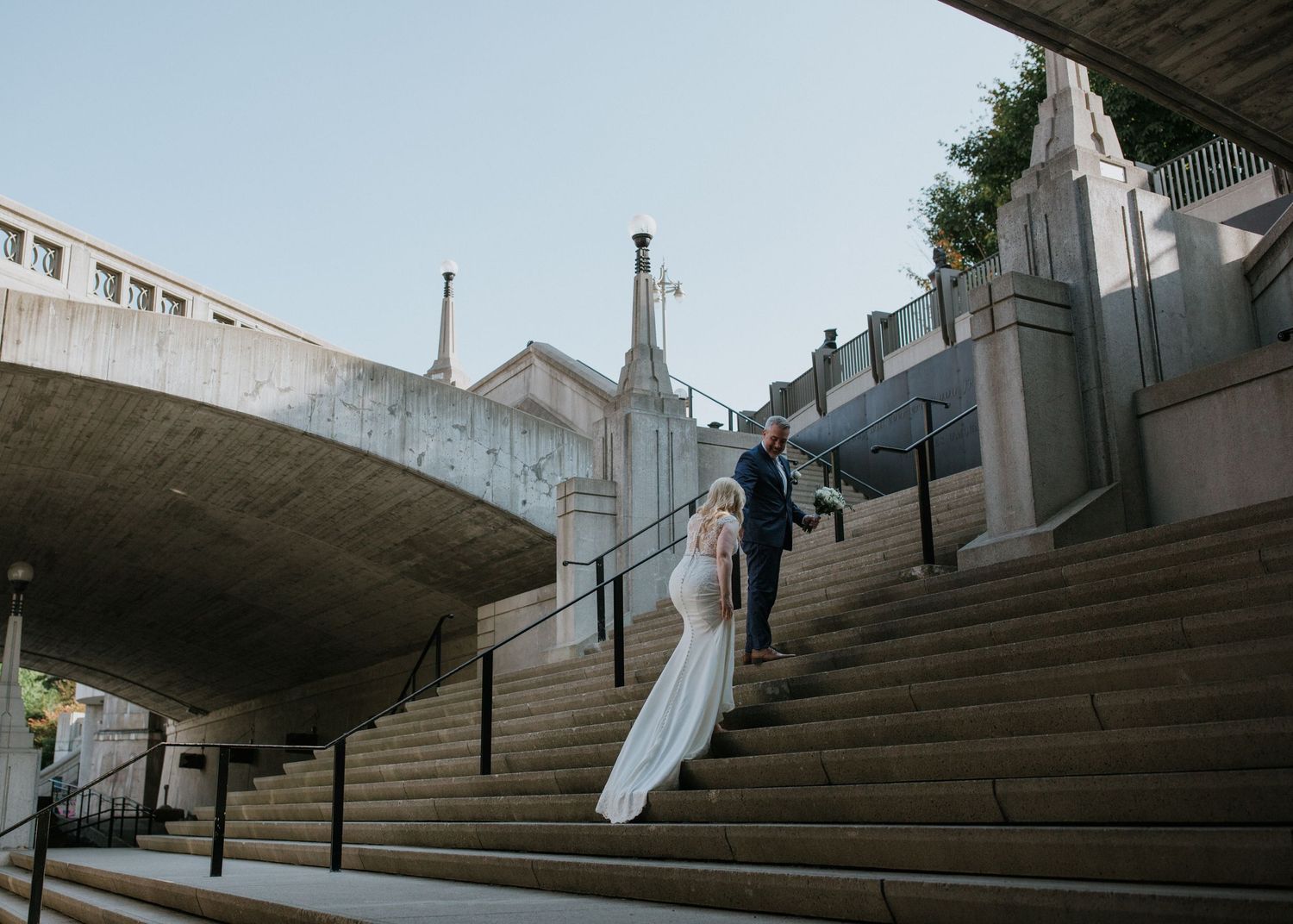 A couple in wedding attire ascends grand concrete steps beneath an architectural overhang.