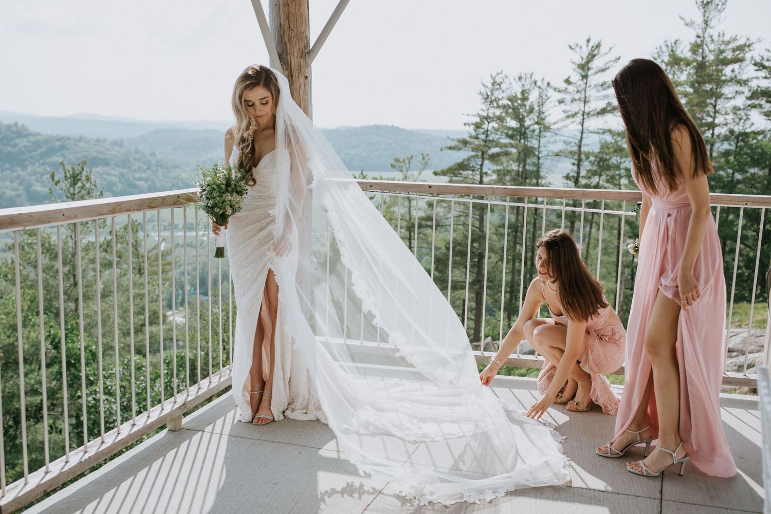 Bride in flowing dress with attendants preparing on mountain view terrace.