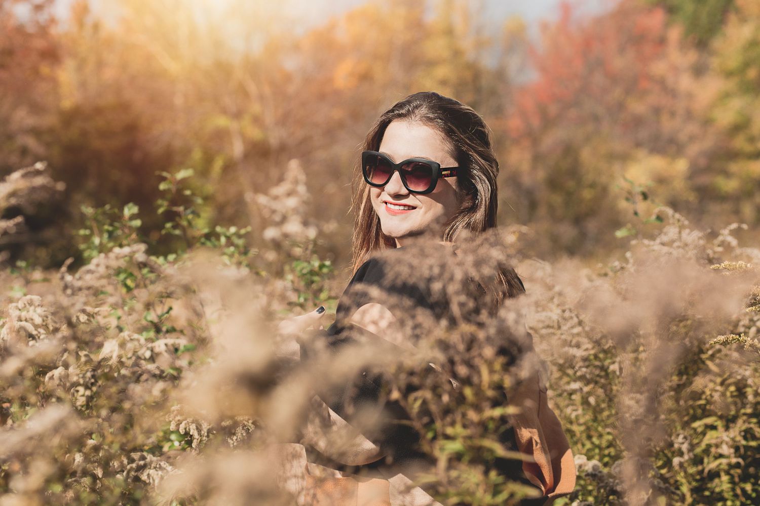 Person wearing sunglasses smiles while surrounded by tall dried grasses at sunset.