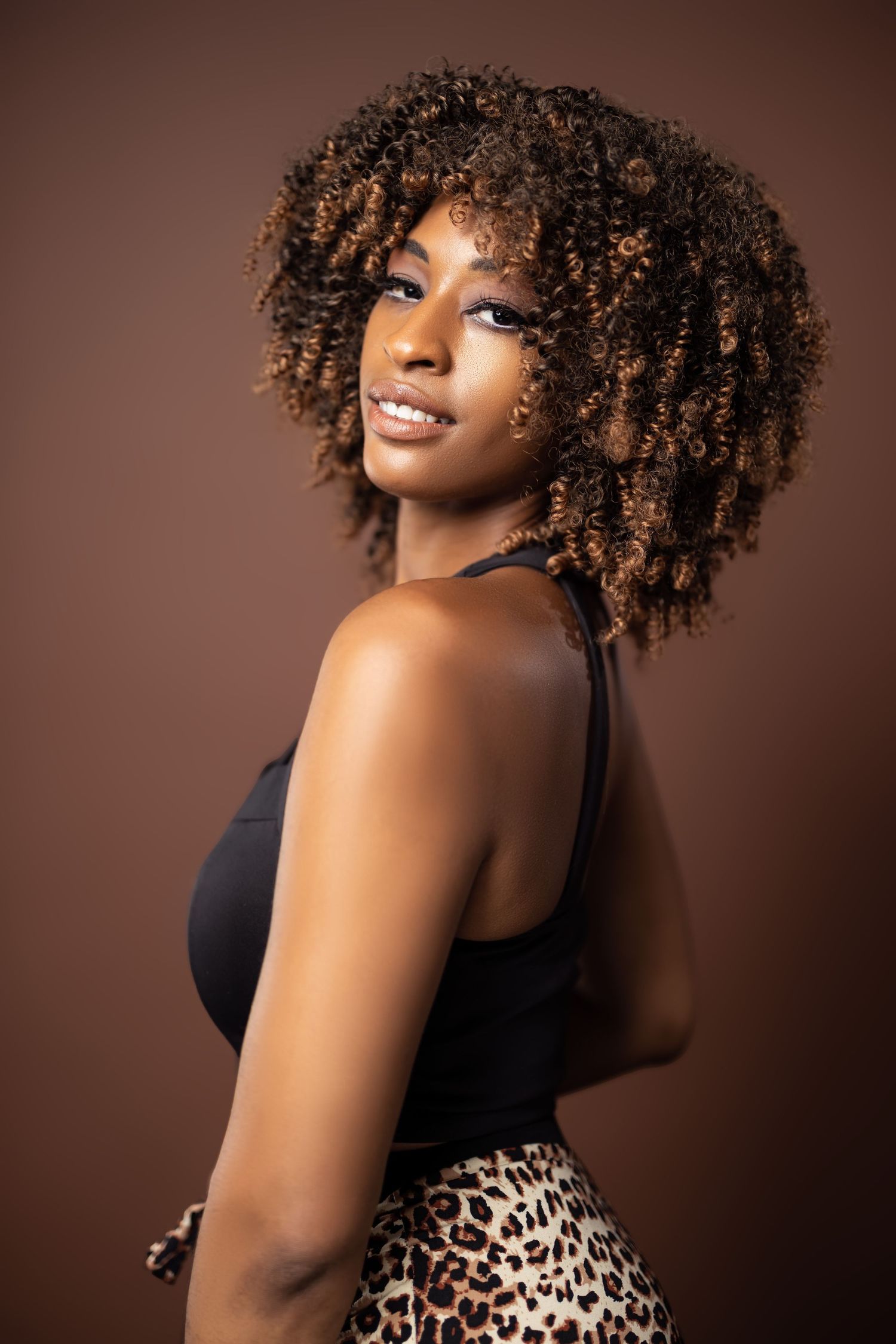 Studio portrait of person with natural curly hair wearing a black top and leopard print skirt against brown background.
