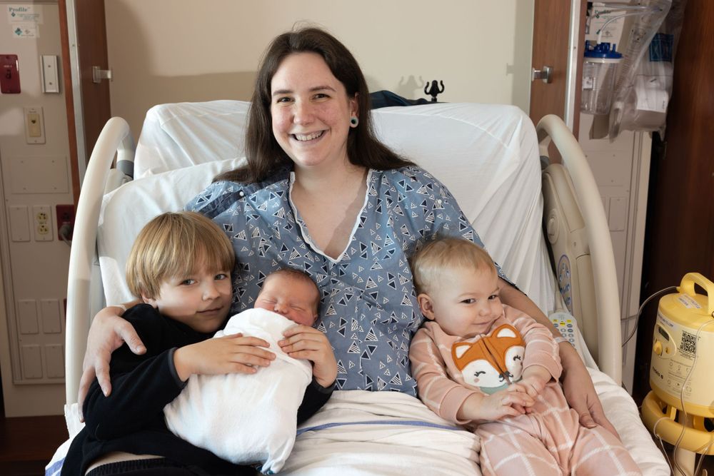 A new mother rests in a hospital bed surrounded by three children.