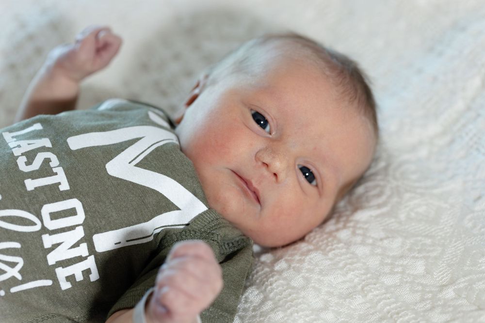 A newborn baby wearing a gray onesie lies on white blanket.