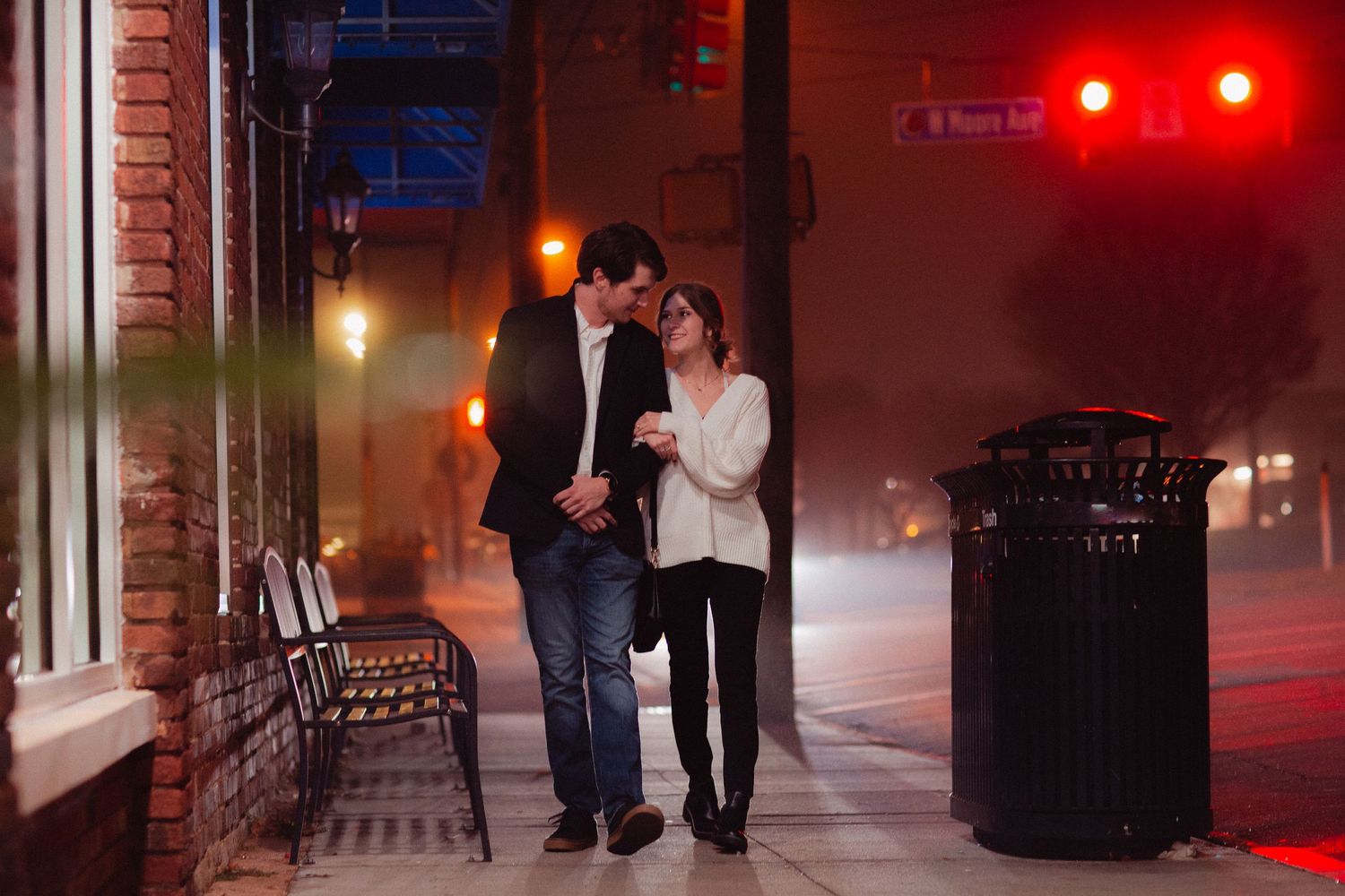 A couple walks together on a city sidewalk illuminated by red lighting at night.