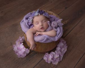 Artistic newborn portrait with purple fabric and floral elements on wooden surface.