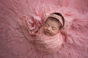 Sweet newborn baby sleeping peacefully wrapped in pink blanket surrounded by soft pink textures.