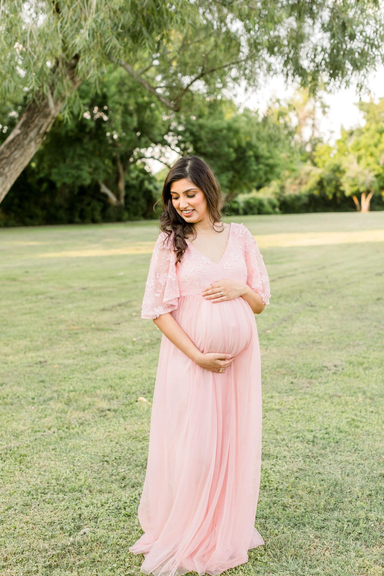 A maternity portrait shows a woman in a flowing pink dress touching her belly in a park setting.