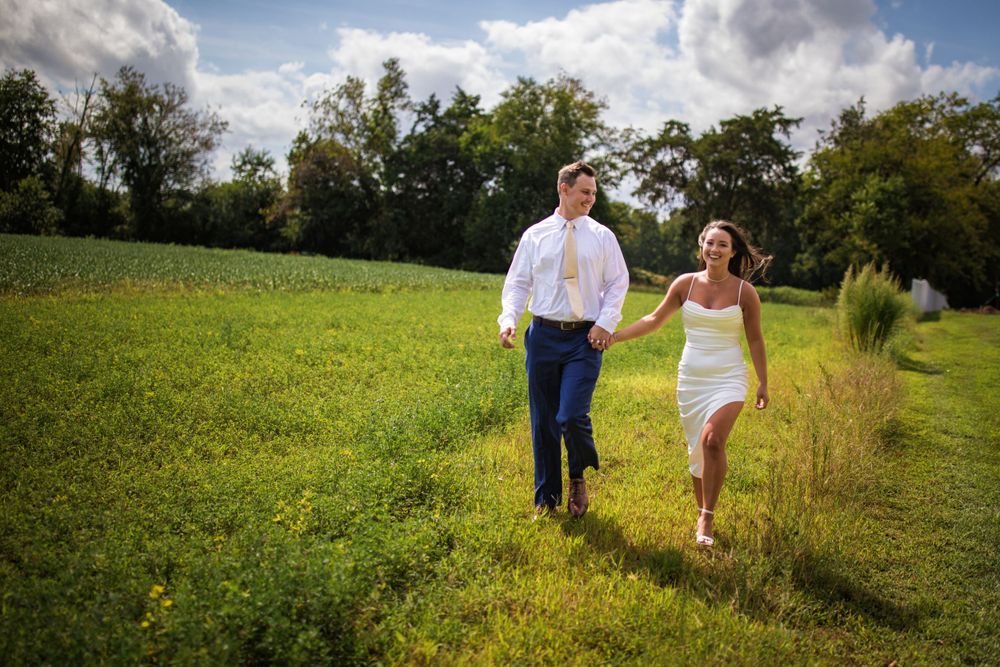 A couple walks hand in hand through a lush green meadow on a summer day with fluffy clouds overhead.