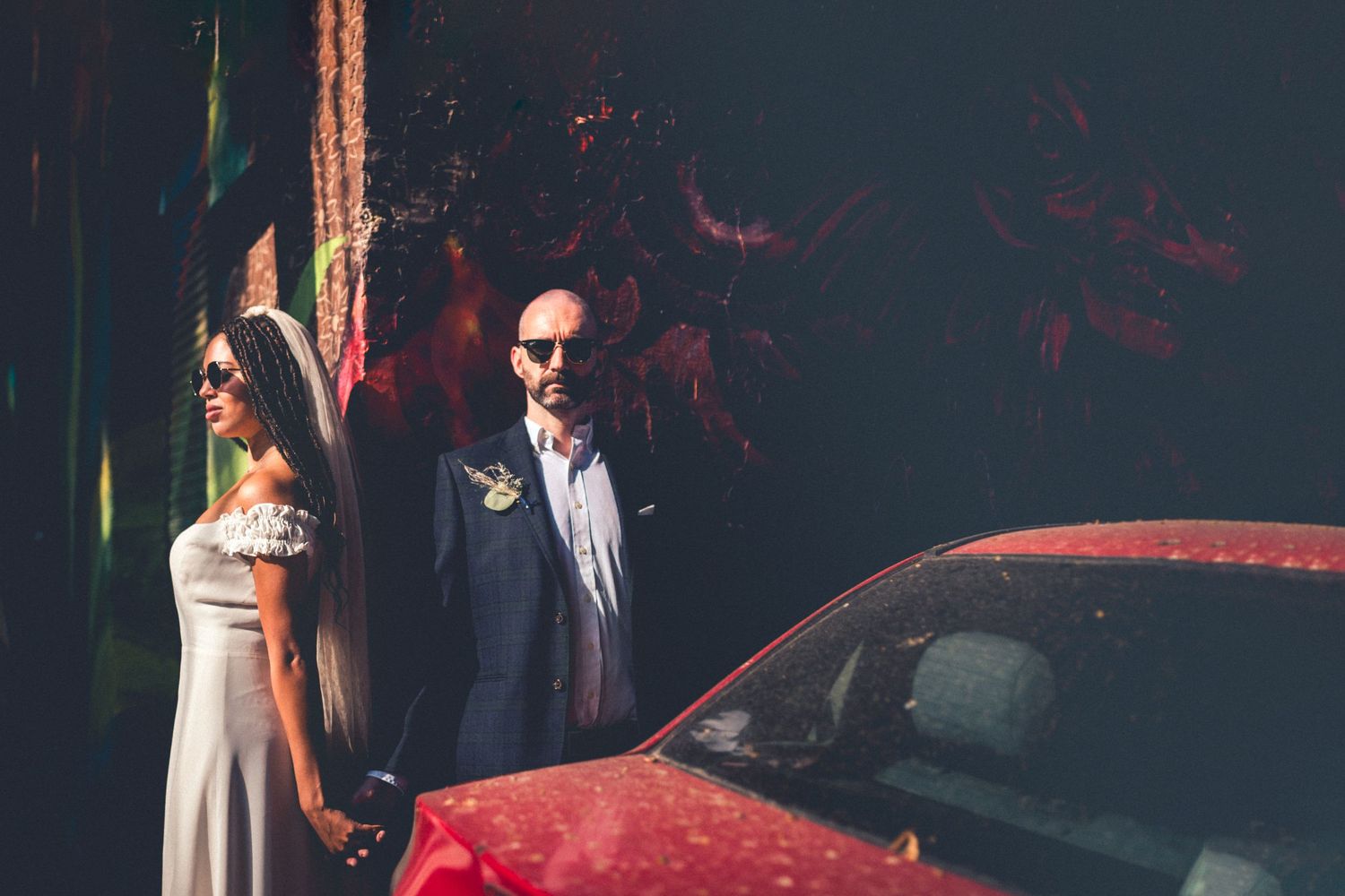 Wedding couple in sunglasses posing beside red car with artistic wall backdrop.