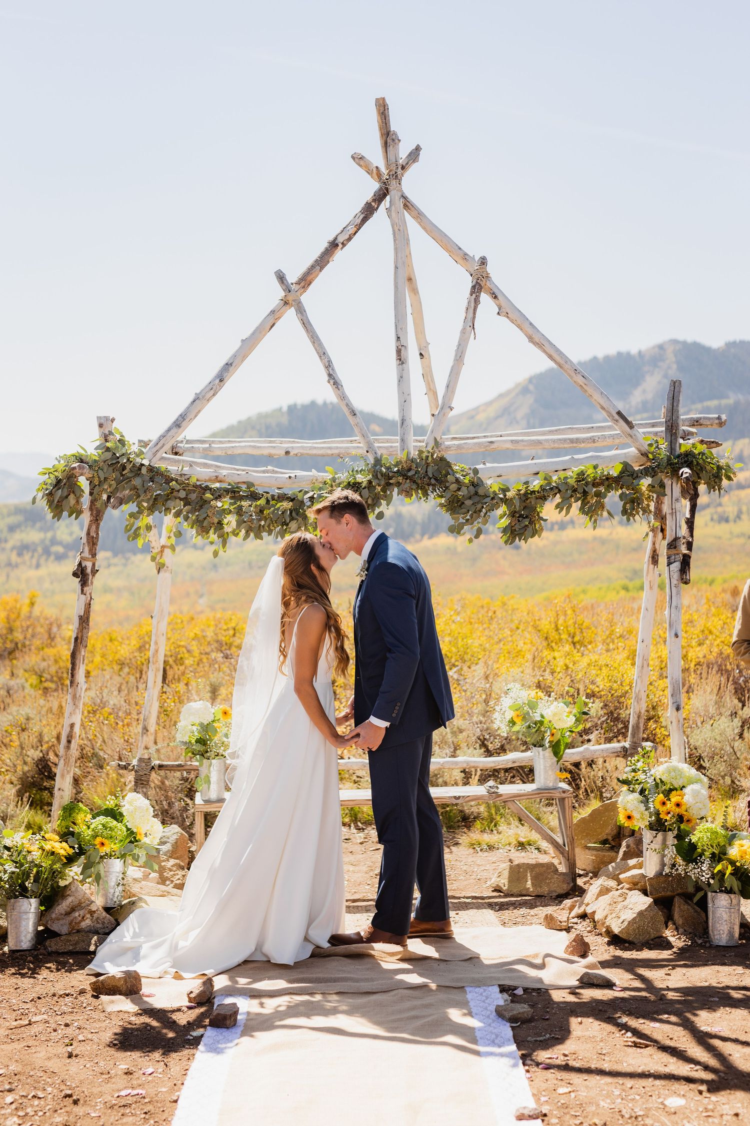 Bride and groom kissing under rustic wooden arch in outdoor fall mountain ceremony.