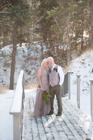 Wedding party in lavender and gray attire pose on snowy wooden deck surrounded by evergreens.
