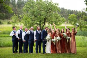 Wedding party poses together on grassy lawn with bridesmaids in rust dresses and groomsmen in navy suits.