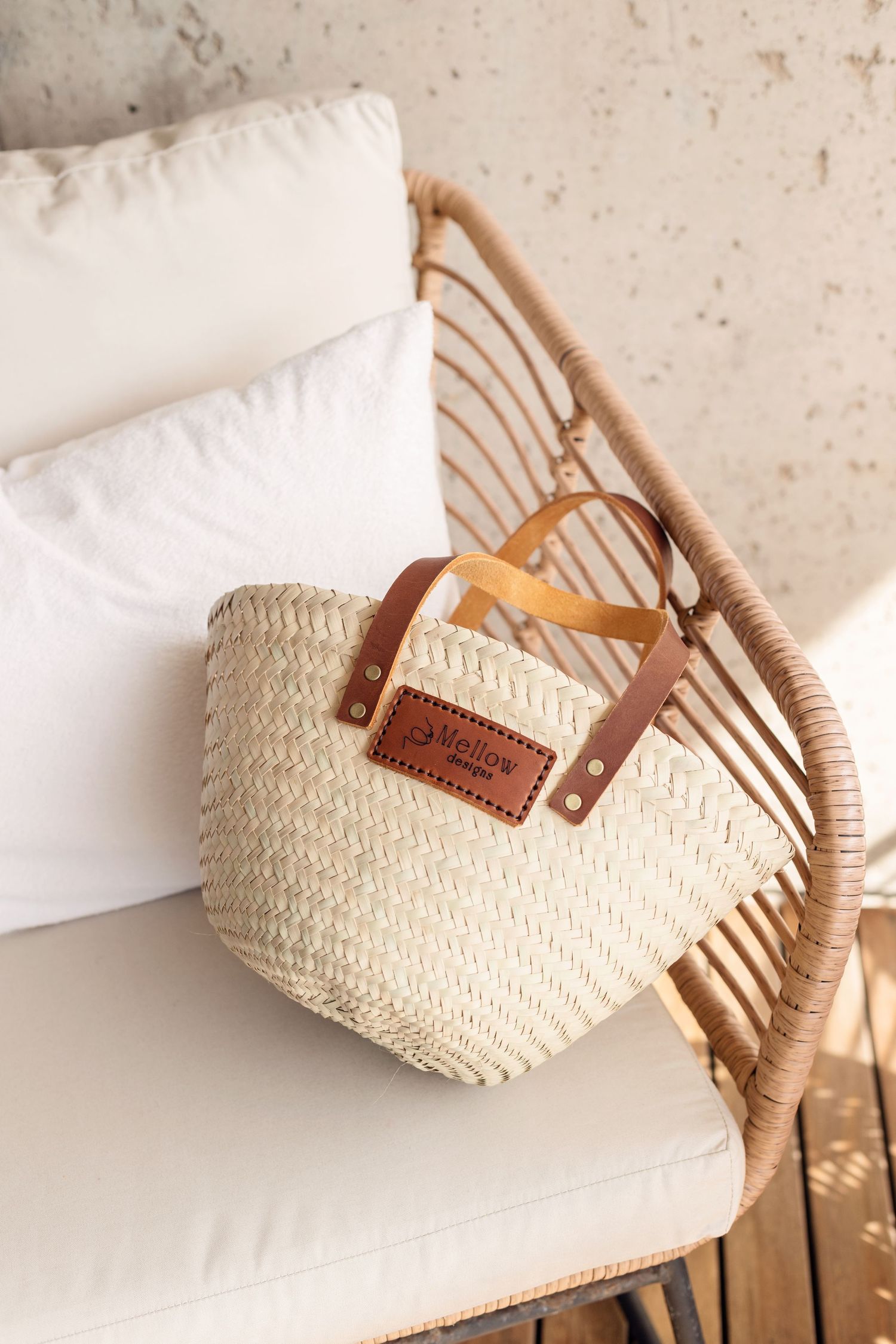 Woven straw market bag with leather handles on rattan chair with white cushions.