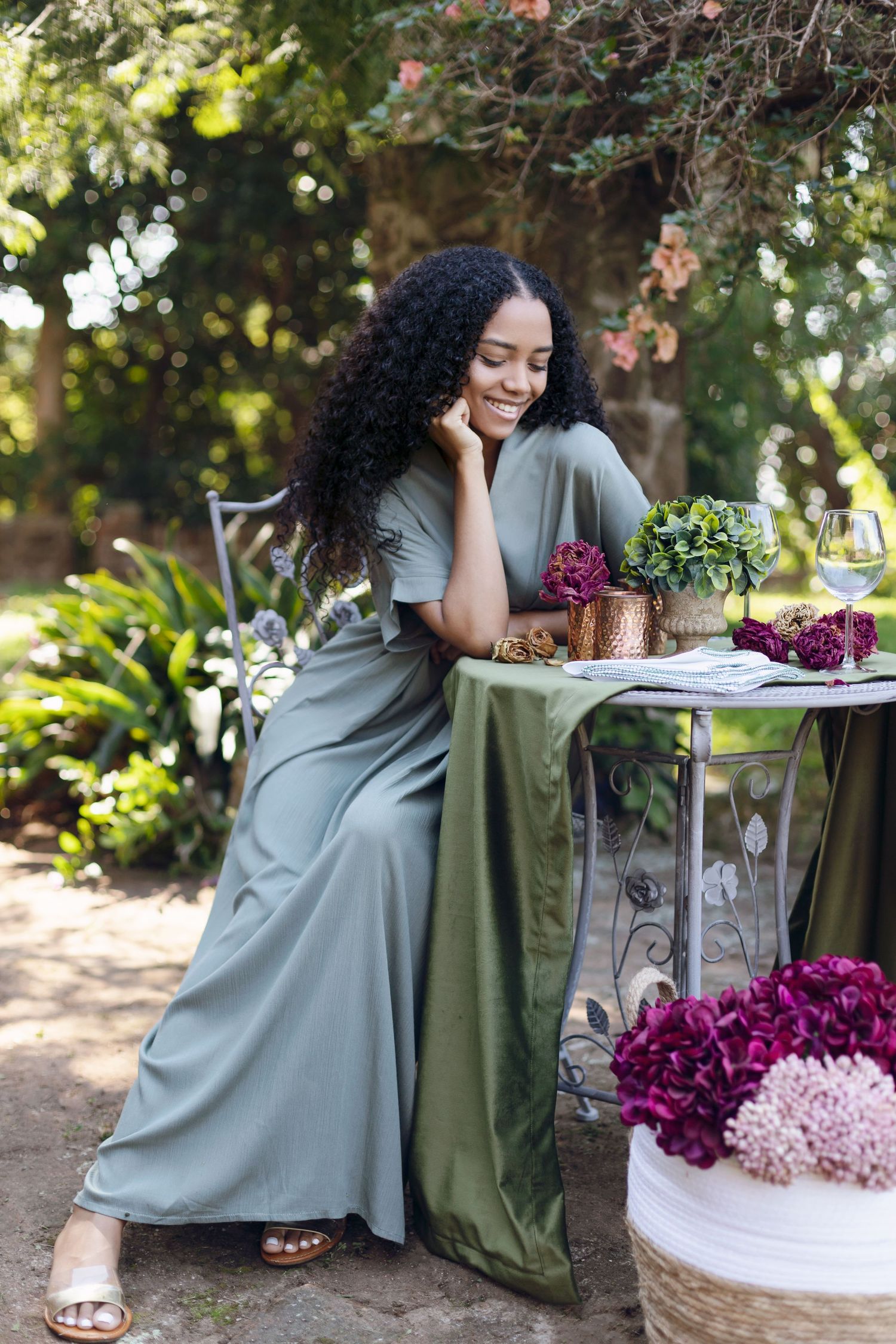 Woman in sage dress sits at garden table surrounded by purple flowers and greenery.