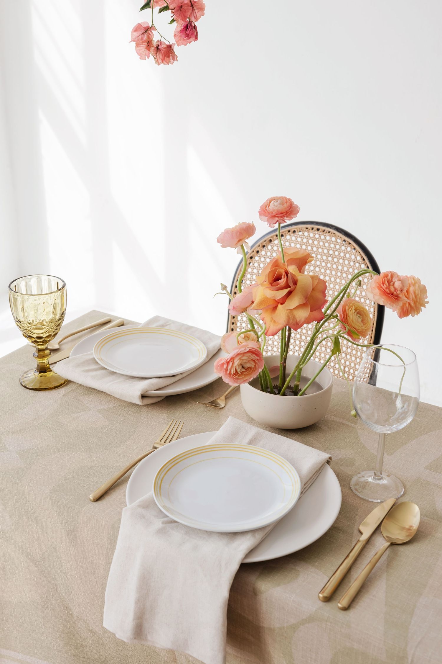 Elegant table setting with coral ranunculus flowers, white plates, gold cutlery and vintage glassware on beige linen.