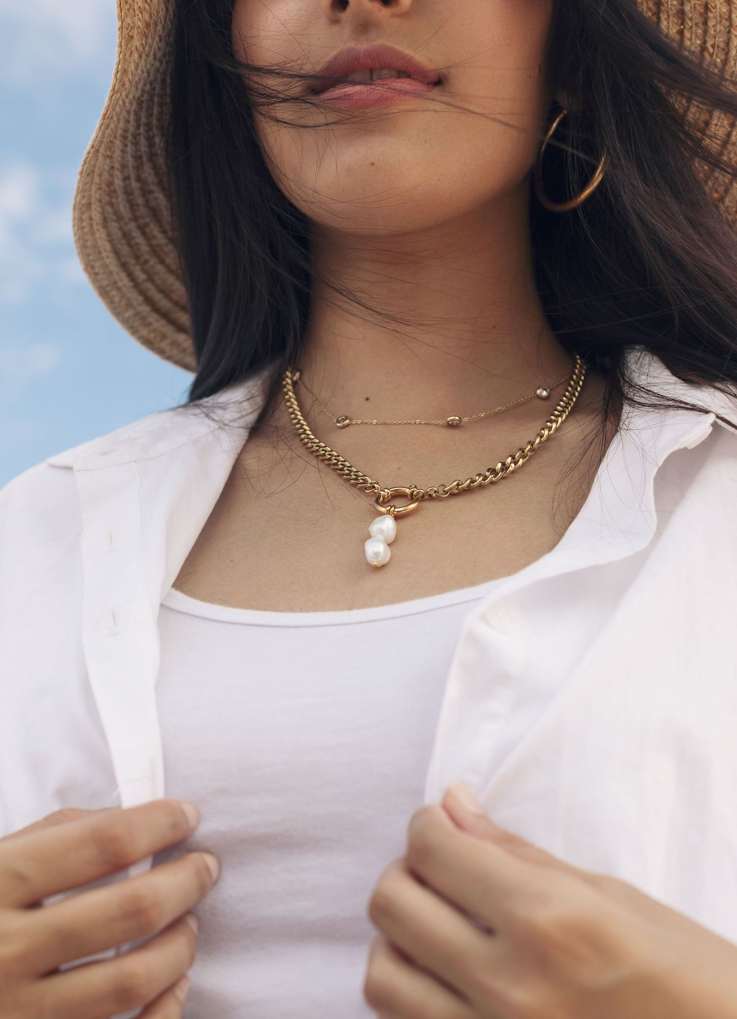 Gold layered necklaces with pearl pendant styled with a white shirt and sun hat.